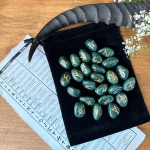 May include: A set of 24 green gemstone runes with gold lettering in a black velvet pouch. The runes are arranged in a circle on the pouch. The runes are used for divination and spell casting.
