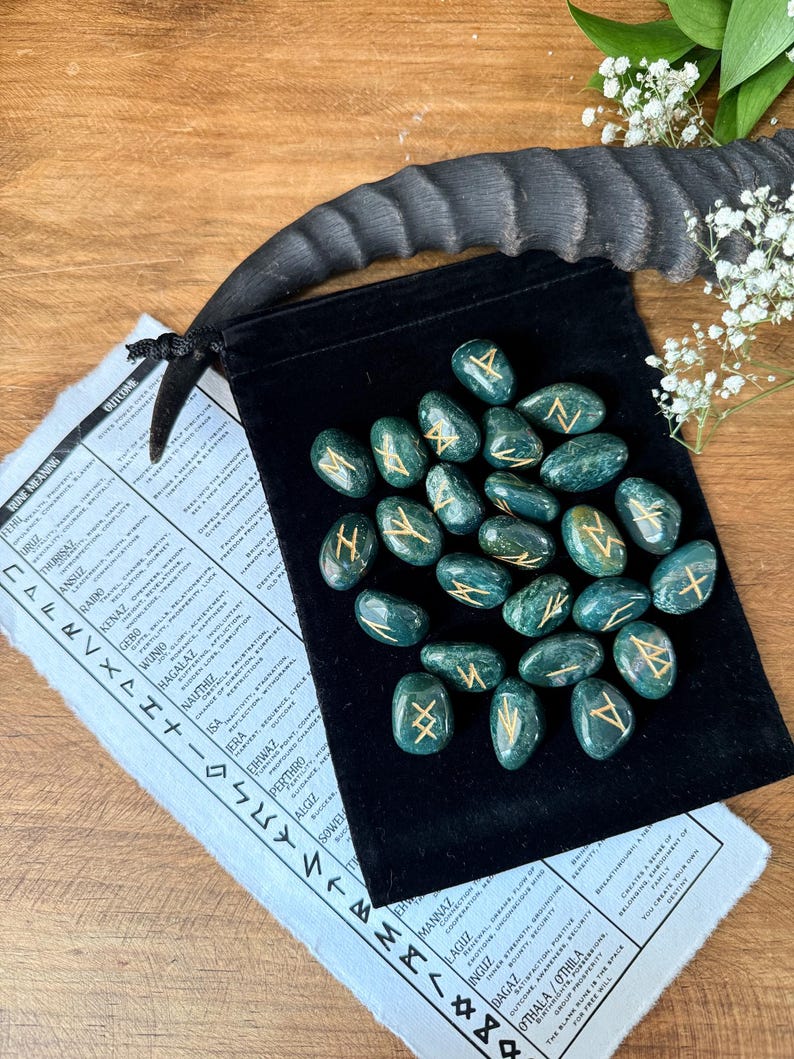 May include: A set of 24 green gemstone runes with gold lettering in a black velvet pouch. The runes are arranged in a circle on the pouch. The runes are used for divination and spell casting.