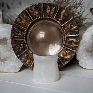 Puede incluir: Una bola de cristal transparente sobre un soporte blanco, rodeada por una rueda zodiacal de color bronce. Cristales de cuarzo blanco a ambos lados. El fondo incluye plantas secas y un recipiente de lat&oacute;n.
