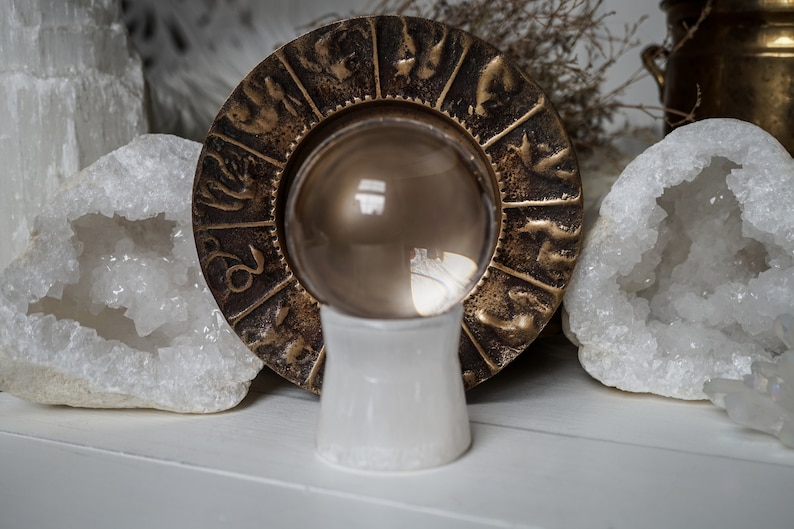 Puede incluir: Una bola de cristal transparente sobre un soporte blanco, rodeada por una rueda zodiacal de color bronce. Cristales de cuarzo blanco a ambos lados. El fondo incluye plantas secas y un recipiente de lat&oacute;n.