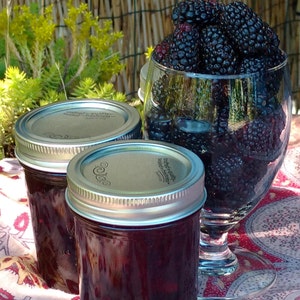 May include: Two glass jars filled with blackberry jam, with a glass of fresh blackberries in the background. The jars have silver lids with text on them.