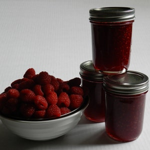 May include: Three glass jars filled with red raspberry jam, with a bowl of fresh raspberries in front. The jars have silver lids.