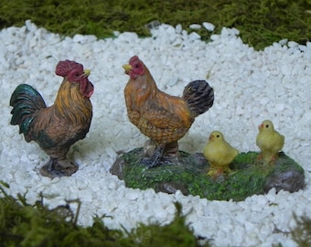 Könnte beinhalten: Eine Miniatur-Bauernhofszene mit zwei Hähnen, einem Huhn und zwei Küken. Die Hähne haben rote Kämme und braune und grüne Federn. Das Huhn und die Küken sind gelb und braun. Die Figuren stehen auf weißen Steinen und grünem Moos.