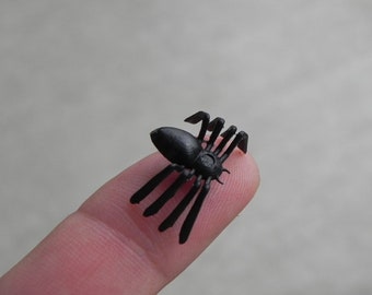 May include: A small, black plastic spider with eight legs and a rounded body. The spider is resting on a fingertip, showcasing its size and detail. The background is blurred, emphasizing the spider.