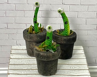 May include: Three miniature potted plants with green stems and googly eyes. The plants are in brown pots and are sitting on a white wooden table.