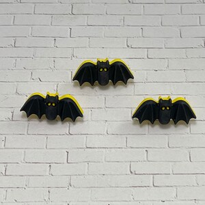 May include: Three black bat-shaped decorations with yellow accents are displayed against a white brick wall. The bats have detailed wings and visible eyes, perfect for Halloween or spooky decor.