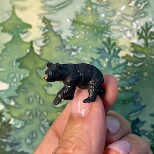 May include: A small, black bear figurine with textured fur detailing and a light-colored snout. The miniature animal is depicted in a walking pose, showcasing a realistic shape and dark coloration throughout its body.