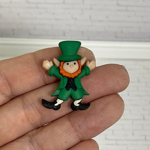 May include: A green leprechaun figurine with a black hat, orange beard, and black shoes. The figurine is holding its arms out to the sides.