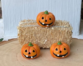 May include: Three orange miniature jack-o'-lanterns with black smiles and green stems are arranged on a wooden slice and a small hay bale. The pumpkins have triangular eyes and white teeth, perfect for Halloween decorations.