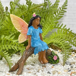 May include: A miniature fairy figurine with a blue dress and peach-colored wings sits on a brown branch. The fairy has black hair and a pink flower in her hair. Green ferns and white pebbles surround the fairy.