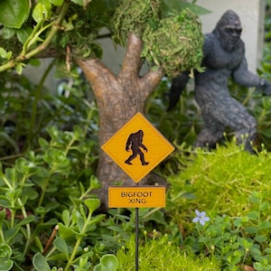 May include: A yellow diamond-shaped sign with a black silhouette of Bigfoot walking, with the text "BIGFOOT XING" on a brown rectangular sign below.
