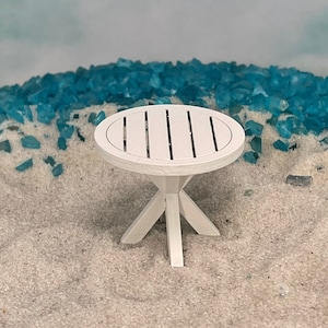 May include: White round table with a slatted top and four legs. The table is sitting on a bed of white sand.