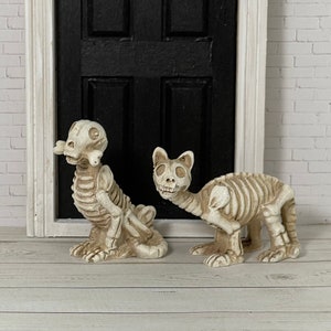 May include: Two miniature skeletal figures of a cat and dog, both in a sitting position. The cat is looking to the right and the dog is looking straight ahead. The figures are made of white painted resin and have a realistic bone structure.