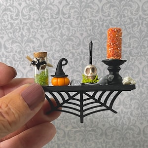 May include: Miniature Halloween-themed shelf with a spiderweb design. It holds a small glass bottle with a bat charm, a pumpkin with a witch hat, a skull, a candle, and a glittery orange candle.