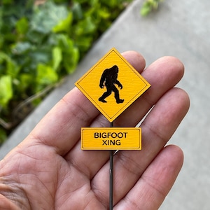 May include: A yellow warning sign with a silhouette of Bigfoot walking, attached to a wooden sign that reads "BIGFOOT XING".