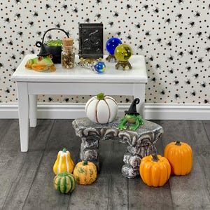 May include: A miniature Halloween-themed display featuring a white table with a cauldron, spell book, and crystal balls. A frog wearing a witch hat sits on a stone bench with pumpkins. The background is a spider web pattern.