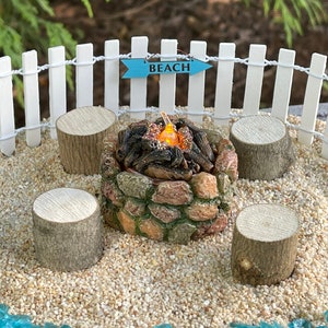 May include: A miniature stone fire pit with a glowing light inside, surrounded by four wooden stumps, on a bed of sand with blue glass stones. A sign above the fire pit reads "Beach".