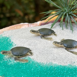 May include: Three small green sea turtle figurines on a white sandy beach with green beads surrounding them.