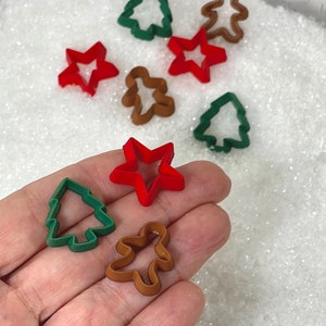 May include: A collection of miniature Christmas-themed cookie cutters in various shapes. The cutters include a green Christmas tree, a red star, and a brown gingerbread man. Additional cutters are scattered in the background. The cutters are small and made of plastic.