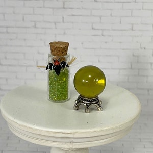 May include: A small glass bottle filled with green beads, topped with a cork and decorative bat. Next to it is a yellow-green crystal ball on a silver stand. The items are on a white, round table.