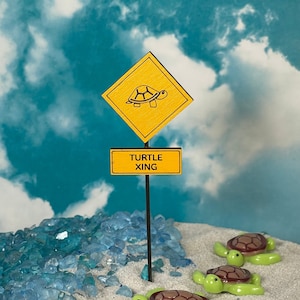 May include: A yellow diamond-shaped sign with a black turtle silhouette and the text "TURTLE XING" on a wooden sign below. Three green turtle figurines are on a white sandy surface with blue glass pieces.