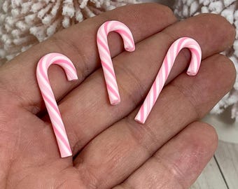 May include: Three miniature candy canes with pink and white stripes. The candy canes are curved and appear to be made of a hard candy material. The candy canes are held in a hand, with a blurred background.