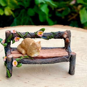 May include: A miniature garden bench made of wood-like material, with a sleeping orange cat figurine. The bench is decorated with pink and yellow flowers and green vines, creating a whimsical scene. Perfect for fairy gardens or miniature displays.