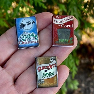 May include: Three miniature Christmas-themed books. The first book is blue with a picture of Santa Claus flying in a sleigh with reindeer. The title is "The Night Before Christmas". The second book is red with a picture of a top hat and the title "A Christmas Carol". The third book is gold with the title "Naughty or Nice".