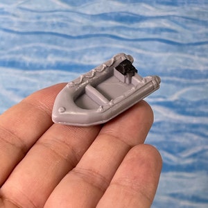 May include: A small, gray toy inflatable boat with a black motor. The boat has a detailed design, including interior seating and exterior trim. The boat is held in a hand against a backdrop of blue water, suggesting a nautical theme.