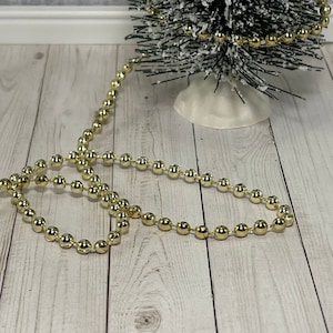 May include: A gold beaded garland with a snowy artificial Christmas tree in the background.