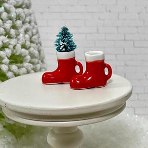 May include: Two miniature red ceramic Christmas boots with white tops sit on a white table. One boot holds a small green Christmas tree. The boots have a handle on the side. A blurred Christmas tree is in the background.