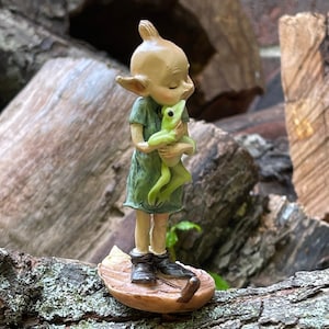 May include: A small, green-skinned figurine of a fairy holding a green frog in its arms. The fairy is wearing a green dress and has closed eyes. The figurine is standing on a leaf-shaped base.