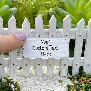 May include: A white picket fence with a small sign that says "Your Custom Text Here". The sign is hanging from the fence with a string.