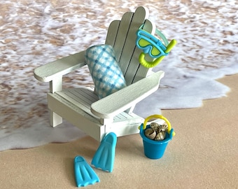 May include: A white wooden beach chair with a blue and white checked pillow, a blue and yellow snorkel and goggles, a blue bucket with seashells, and two blue flippers.