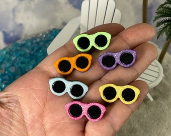 May include: Six pairs of miniature sunglasses in various colours, including green, orange, purple, blue, yellow, and pink. The sunglasses are made of glitter-covered wood and have black lenses.