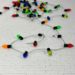 May include: A string of miniature Christmas lights with red, green, blue, yellow, and orange bulbs. The lights are strung together with clear wire and are hanging on a white brick wall.