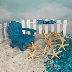 May include: A miniature beach scene with a blue Adirondack chair, white picket fence, and a sign that says "BEACH". Several tan starfish are scattered on the sand, some with pearl accents. Blue glass pieces add a water effect.
