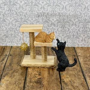 May include: A wooden cat tree with two levels and a dangling toy. A brown cat is sitting on the top level and a black cat is standing on the bottom level.