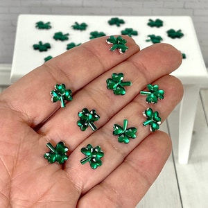 May include: A collection of green, jewel-toned shamrock embellishments. The four-leaf clovers are held in a hand, with more scattered on a white surface in the background. The shamrocks have a metallic silver outline.