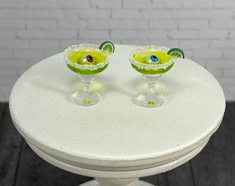 May include: Two miniature margarita glasses with green liquid and a gummy eyeball inside each. The glasses are rimmed with white sugar and have a lime wedge garnish.