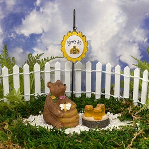 May include: A miniature brown bear figurine holding a honey pot with a bee on it. The bear is wearing a flower garland. There is a yellow sign that says "Honey $5" and three miniature honey jars on a wooden log.