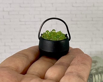 May include: A miniature black cauldron with a curved handle, filled with green, sparkling beads. The cauldron is small enough to fit in the palm of a hand, suggesting a decorative or novelty item. The background is a white brick wall.