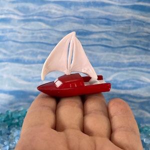 May include: A small red toy sailboat with a white sail. The sailboat is sitting on a hand.