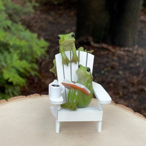 May include: Two green frogs sitting on a white adirondack chair. One frog is reading a book and the other is looking at the viewer. There is a white coffee cup on the chair's armrest.