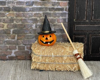 May include: A small orange pumpkin wearing a black witch hat sits on a bale of hay with a wooden broom leaning against it.