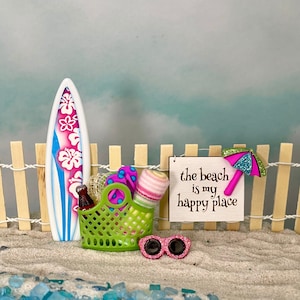 May include: A miniature beach scene with a surfboard, a green basket, a pink and blue striped towel, a pink and blue beach umbrella, and a sign that reads "the beach is my happy place".