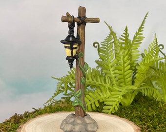 May include: A miniature wooden lamp post with a black lantern and a vine growing up the side. The lamp post is on a wooden base with a gray stone base.
