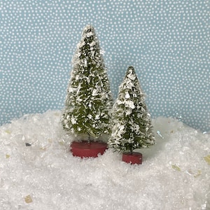 May include: Two miniature green Christmas trees with white snow-like accents and red bases. The trees are set in a bed of white, sparkly material against a light blue background with white dots.