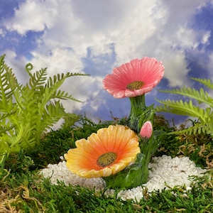 May include: A miniature garden ornament featuring two colorful flowers. One flower is pink, the other is yellow and orange. The flowers are set against a backdrop of green moss, ferns, and a blue sky with clouds.