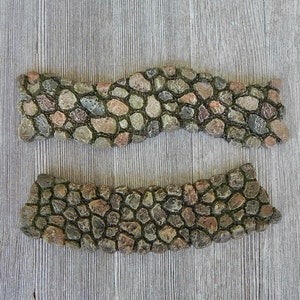 May include: Two pieces of decorative stone edging for a garden path or walkway. The edging is made of small, rounded stones in shades of brown, gray, and green. The stones are arranged in a wavy pattern.